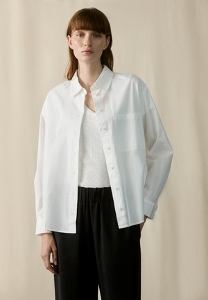 Chemise en coton blanc à coupe décontractée, avec un devant à boutons et une poche poitrine, associée à un pantalon noir à taille élastique. Texture lisse, manches longues.