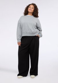 Felpa grigia corta con polsini a costine, abbinata a pantaloni larghi neri. Il modello indossa scarpe slip-on color crema. Dettaglio del logo semplice sulla maglietta.