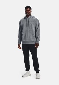 Grå hoodie med frontlomme og snøre, med et hvidt logo. Parret med sorte sweatpants og hvide sportssko.