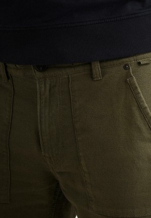Close-up van olijfgroene broek met gedetailleerd stiksel, riemlussen, voorzakken en een zwarte geribde tailleband bovenaan.