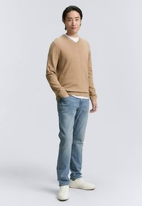 Vaaleanruskea v-kaula-aukkoinen pusero, valkoinen aluspaita, siniset farkut ja valkoiset tennarit. Kapealinjainen, pehmeä tekstuuri ja rento muotoilu. Seisova asento.