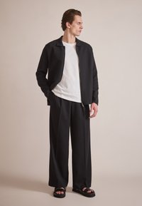 Svart zip-up jakke over en hvit T-skjorte, kombinert med løse svarte bukser og svarte sandaler. Enkelt design, jevn tekstur, uformell stil.