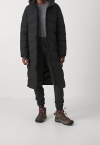 Dlouhý černý péřový kabát s kapucí, s prošívaným vzorem, předním zipem a oranžovými detaily. Kombinovaný s tmavými kalhotami a trekovými botami.