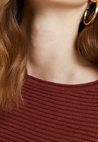 Haut côtelé couleur rouille avec un col rond. Cheveux longs et ondulés et une grande boucle d'oreille dorée sont visibles, mettant en valeur la texture et la brillance.