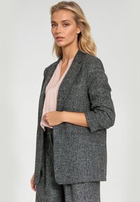 Szary żakiet o fakturowanym wykończeniu z dopasowanym krojem, zapinany na jeden guzik, z rękawami do łokci, w połączeniu z jasnoróżową bluzką pod spodem.