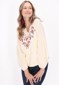Donna che indossa una blusa crema con ricami floreali rossi e blu e maniche lunghe a palloncino, abbinata a jeans scuri, sorridente e con uno sguardo di lato.