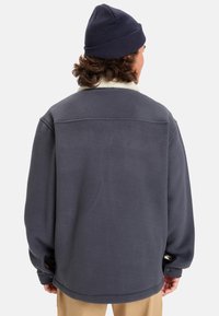 Tummanharmaa fleece-takki, jossa on valkoinen kaulus. Takissa on nappisuljenta ja sivuhalkiot. Pehmeä materiaali ja rento istuvuus.