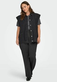 Zwarte denimmouwloze vest met afgeronde schouders, zilveren drukknopen, twee voorzakken, gedragen over een grijs T-shirt en bijpassende broek.