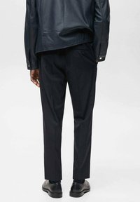 Schwarze maßgeschneiderte Hose mit geradem Schnitt, aus glattem Material. Kombiniert mit schwarzen Lederschuhen und einer taillierten schwarzen Lederjacke.