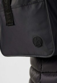 Borsa nera in nylon con superficie liscia, dotata di una tasca frontale e un logo JOTT rotondo in gomma nera. Include un manico testurizzato.