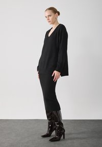 Svart strikket genser med V-hals, kombinert med en rippet svart skjørt og glinsende, knelange stiletthæler, presentert i en minimalistisk setting.