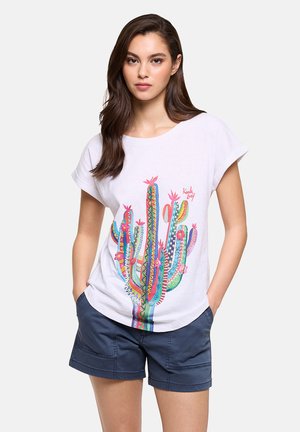 Joven con cabello largo y castaño, vistiendo una camiseta blanca con estampado colorido de cactus y pantalones cortos azul marino, de pie con las manos en los bolsillos.