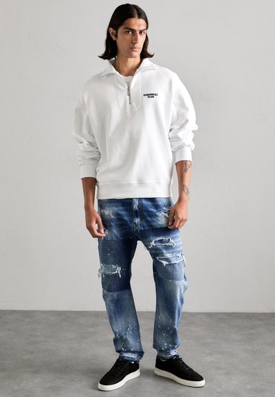 Witte zip-up sweatshirt met een ontspannen pasvorm en geribbelde manchetten, gecombineerd met distressed blauwe jeans met gepatchte gebieden en verfpots, zwarte sneakers.