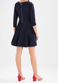 Mörkblå knälång klänning med trekvartsärm, utsvängd kjol och en guldfärgad dragkedja bak. Bärs med röda blockklackade sandaler.