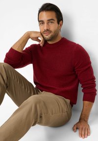 Maglione rosso lavorato a maglia con texture a coste e collo a girocollo, abbinato a pantaloni beige. Il modello è seduto con una mano sul mento.