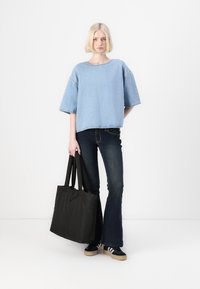 Top oversize in denim blu chiaro con maniche corte, abbinato a jeans scuri a zampa d'elefante. Borsa a tracolla nera e sneakers nere con strisce bianche.