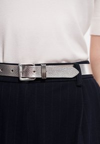 Ceinture en cuir argenté brillant avec une boucle rectangulaire, fixée sur un pantalon à rayures sombres, portée avec un haut blanc à manches courtes.