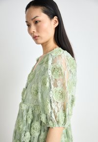 Robe en dentelle florale vert menthe avec des manches trois-quarts, des appliqués en rose texturé et une coupe décontractée avec un décolleté en V.
