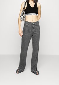 Top corto in velluto nero con un logo bianco di Tommy Jeans, abbinato a jeans in denim grigio a vita alta e calzature nere con dettagli arancioni.