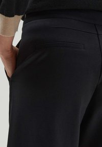 Black pants made of smooth fabric, featuring a back pocket with stitched detail. The waistband is slim, and texture appears slightly stretchy.