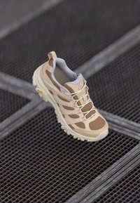 Beige idrettssko med overdel i mesh og semsket skinn, polstret krage, teksturert såle og kontrasterende brune detaljer. Snøring og løftekabel inkludert.