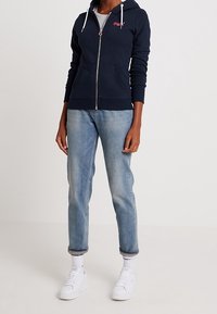 Felpa con zip di colore blu navy, dotata di cappuccio e tasche frontali, abbinata a jeans tapered di un blu chiaro e scarpe da ginnastica bianche. Tessuto con trama e cordini.