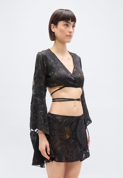 Γυναίκα που φοράει μαύρο διάφανο crop top με μακριά μανίκια και ταιριαστή φούστα με περίπλοκη κεντητή λουλουδάτη διακόσμηση και λεπτομέρειες δέσιμο περιμετρικά.