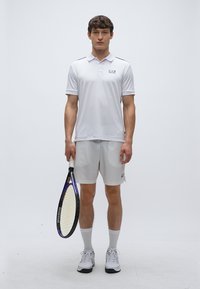 Biele polo tričko s krátkymi rukávmi a čiernymi prvkami, skombinované so svetlosivými šortkami. Model drží tenisovú raketu. Biele športové topánky a ponožky.