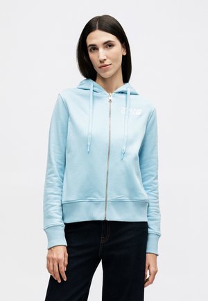 Γυναίκα με ίσια σκούρα μαλλιά, ντυμένη με ανοιχτό γαλάζιο φούτερ με φερμούαρ και σκούρο παντελόνι, που στέκεται μπροστά σε ένα απλό άσπρο φόντο.