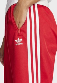 Hand in de ritssluiting van rode Adidas broek met witte drie strepen en het Adidas trefoil-logo aan de zijkant.