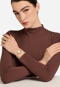 Bracciale d'argento con un ciondolo circolare, indossato su un polso. Maglia a maniche lunghe marrone. Tessuto di texture liscia, design minimalista. Sfondo bianco.