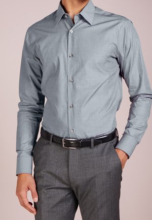 Mann trägt hellblaues Hemd, in graue Anzughose mit schwarzem Ledergürtel gesteckt, eine Hand in der Hosentasche.