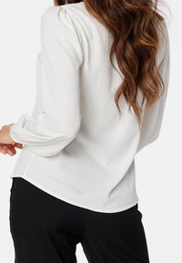 Biała bluzka z delikatnie marszczonym ramieniem, długimi rękawami i zaokraglonym dołem. Wykonana z lekkiego materiału o gładkiej teksturze, w połączeniu z czarnymi spodniami.