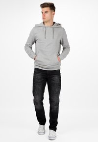 Felpa grigia con cappuccio e cordini, abbinata a jeans neri strappati e sneakers bianche, in piedi contro uno sfondo semplice.