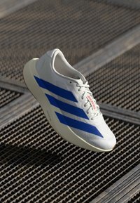 Baskets de sport blanches avec des rayures diagonales bleues et un texte "ADIZERO" rouge, présentant un dessus texturé et un design de semelle léger.