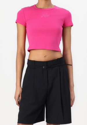 Kvinde iført en lys pink kort T-shirt med teksten "Hago" og højtaljede sorte shorts med bælte mod en ensfarvet baggrund.
