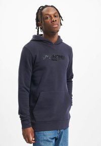 Ung mand med flettet hår iført en marineblå hættetrøje med teksten "JACK & JONES PREMIUM" og blå jeans, stående mod en simpel hvid baggrund.