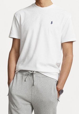 Mand iført en enkel hvid t-shirt med korte ærmer, et lille blåt logo og lysegrå joggingbukser med hænderne i lommerne.