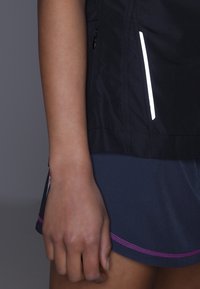 Czarna sportowa kurtka z odblaskowymi akcentami, w połączeniu z ciemnoniebieskimi szortami z różowym wykończeniem. Materiał jest gładki i lekki.