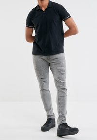 Chemise polo noire avec des accents blancs, assortie à un jean gris clair distressed et des baskets montantes noires. Se tenant sur un fond uni.