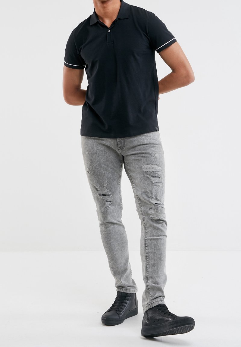 Chemise polo noire avec des accents blancs, assortie à un jean gris clair distressed et des baskets montantes noires. Se tenant sur un fond uni.