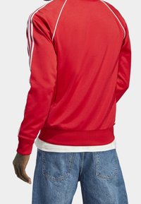 Červená sportovní bunda s bílými pruhy podél ramen, vyrobená z hladké látky. Kombinováno s světle modrými džínami.