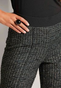 Tweed broeken met een zwarte tailleband, voorzien van een textuurpatroon van grijze en blauwe draden; een grote zwarte ring accentueert de hand.