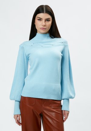 Lichtblauwe trui met hoge nek, textuur in de mouwen en een geribbelde zoom. Heeft decoratieve details over de borst. In combinatie met een bruine broek.