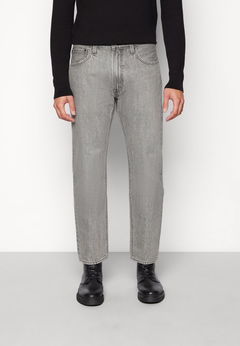 Homme portant un jean droit gris clair, des bottes noires à lacets et une chemise noire à manches longues, debout contre un mur blanc.
