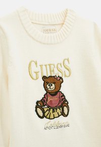 Fehér pulóver, amelyen medve mintázat található, rózsaszín felsővel és szoknyával. A medve fölött arany színnel hímzett "GUESS" felirat. Bordás nyakkivágás.