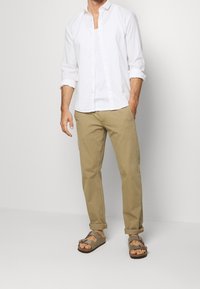 Camicia bianca a maniche lunghe, chinos beige e sandali marroni con doppie cinghie. Look casual con maniche arrotolate e pantaloni alla caviglia.