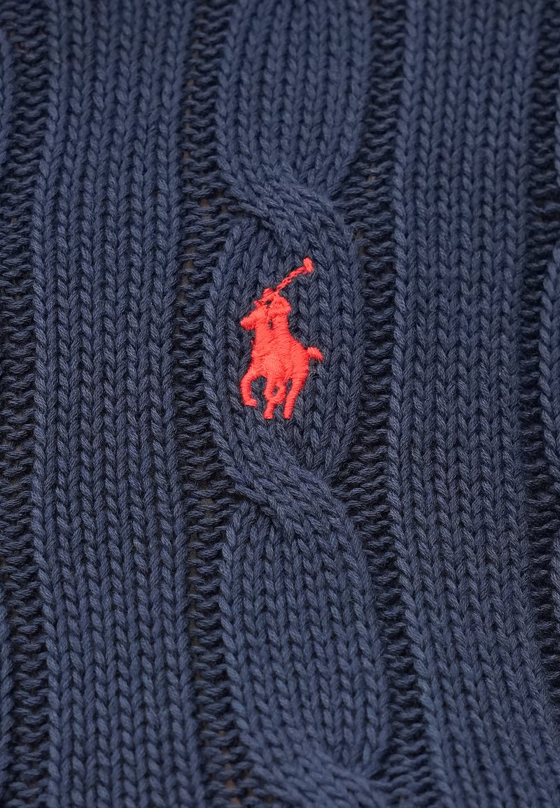 Pull en tricot bleu marine avec un motif en câble, orné d'un logo rouge brodé représentant un cheval et un cavalier sur le devant.