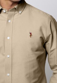 Beige Hemd mit Knöpfen und Kragen, das ein kleines gesticktes Design auf der linken Brust aufweist. Hergestellt aus glattem Stoff mit taillierter Silhouette.