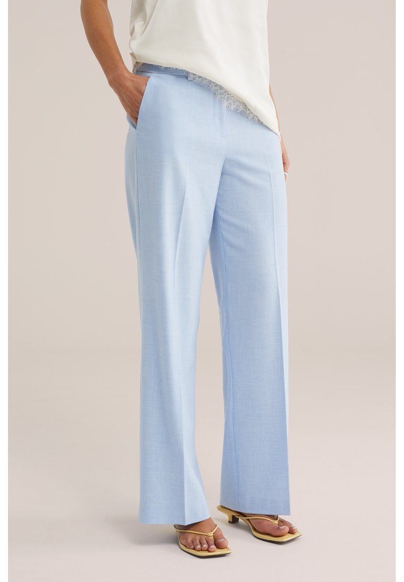 Person trägt hellblaue, weit geschnittene Hosen mit Seitentaschen, ein weißes Oberteil mit Spitzenabschluss und gelbe Sandalen mit quadratischer Zehenpartie und Absatz.